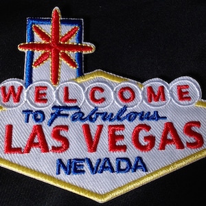 Puede incluir: Un parche blanco en forma de diamante con un contorno amarillo. El parche presenta el icónico letrero "Welcome to Fabulous Las Vegas, Nevada" con una estrella roja en la parte superior. El texto está bordado en rojo y azul.