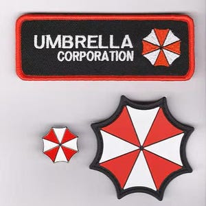 May include: A rectangular black embroidered patch with a red border, featuring the white text UMBRELLA CORPORATION and a red and white umbrella logo. Below are two additional umbrella-shaped patches in red and white with black borders.