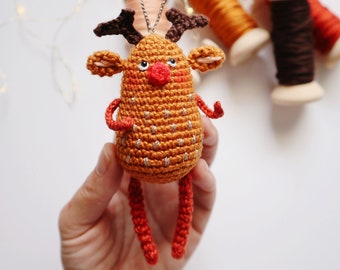 May include: A crocheted reindeer ornament with a brown body, red nose, and brown antlers. The ornament is hanging from a silver string and is being held by a hand.