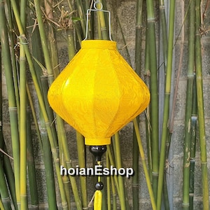 May include: A yellow silk lantern with a tassel hanging from the bottom. The lantern is hanging from a metal hook against a backdrop of bamboo.