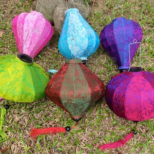 May include: Six vibrant silk lanterns in pink, blue, green, red, and purple sit on grass. Each lantern has a distinct shape and a decorative tassel. The lanterns have a metal handle for hanging, perfect for outdoor decor.