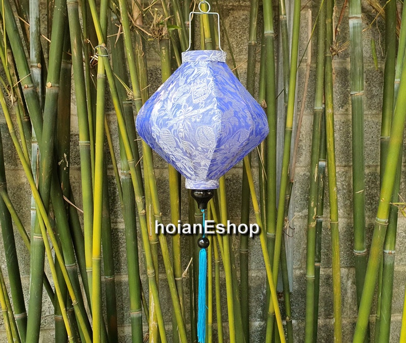 May include: A blue and white silk lantern with a tassel hanging from the bottom. The lantern is hanging from a hook in front of a bamboo wall.