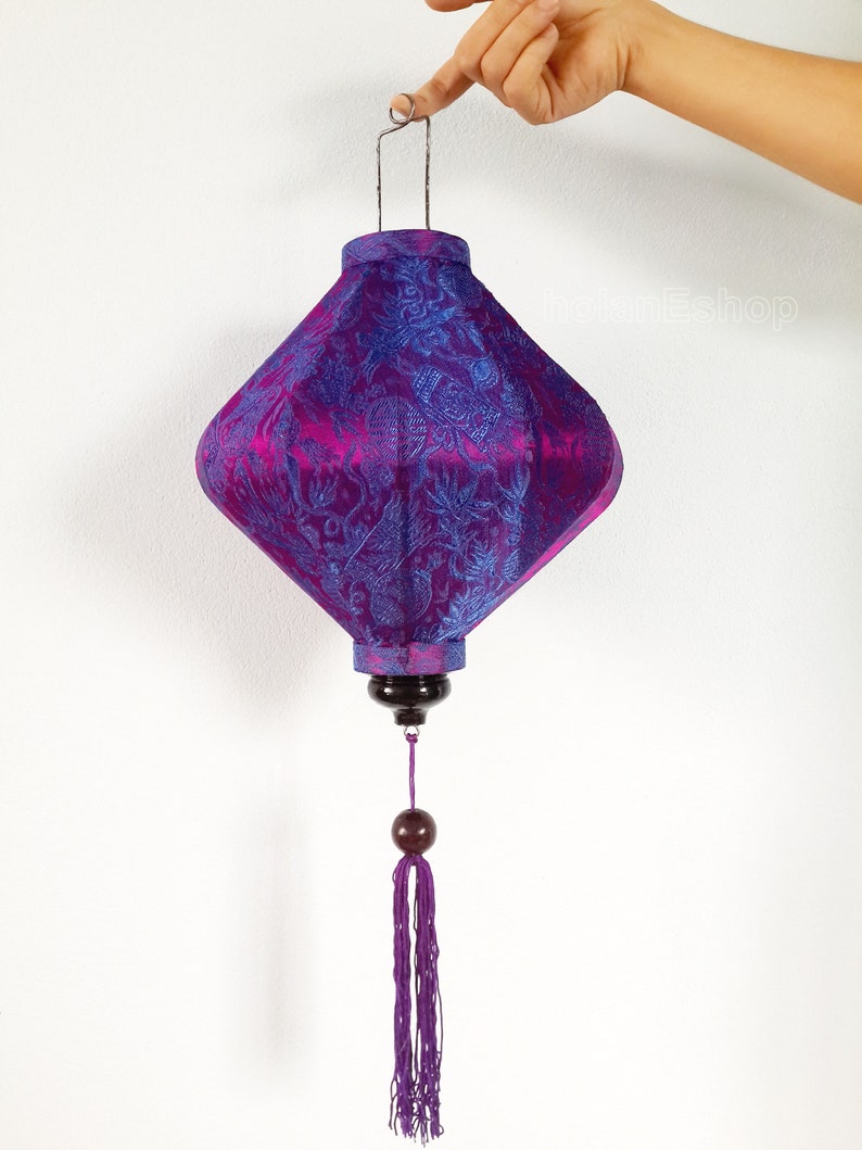 May include: A purple and blue silk lantern with a floral pattern and a tassel. The lantern is hanging from a metal hook.