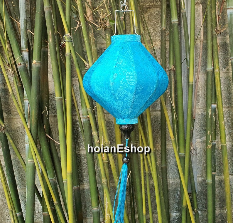 May include: A blue silk lantern with a tassel hanging from the bottom. The lantern has a white design and is hanging from a bamboo background.