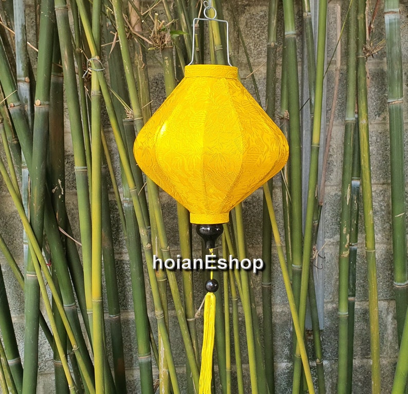 May include: A yellow silk lantern with a tassel hanging from the bottom. The lantern is hanging from a metal hook against a backdrop of bamboo.