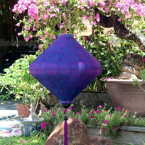 May include: A purple and blue silk lantern with a tassel hanging from a tree branch. The lantern is decorated with a floral pattern and is hanging from a metal hook.