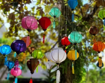 May include: A collection of colorful paper lanterns hanging from a tree branch. The lanterns are in various shapes and sizes, including round, square, and rectangular. Some of the lanterns are decorated with patterns, while others are plain. The lanterns are all lit up, creating a festive atmosphere.