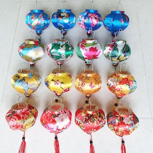 May include: A collection of colorful, silk Chinese lanterns. The lanterns feature floral and nature-inspired designs in shades of blue, green, yellow, and red. Each lantern has a tassel and a metal hanging loop.