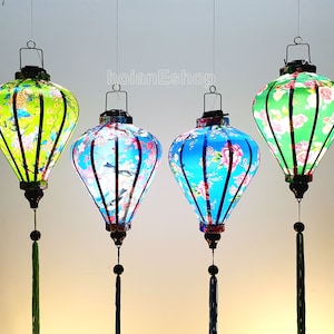 May include: Four hanging lanterns with floral patterns in green, blue, and pink. Each lantern has a black top and a tassel. The lanterns are lit from within, creating a soft glow. The text "hoianEshop" is visible.