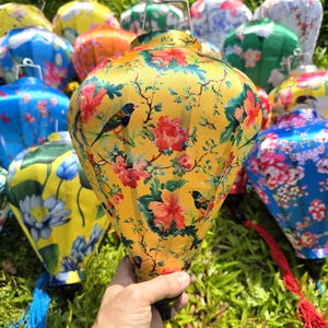 May include: A collection of colorful, patterned paper lanterns. The central lantern is yellow with a floral and bird design. Other lanterns are blue, green, and orange, with various floral patterns. Each lantern has a metal hanger and a tassel.