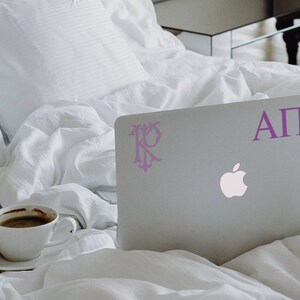 Könnte beinhalten: Ein silberner Laptop mit einem violetten Aufkleber, auf dem "ΑΠΦ" steht, und einem weiteren Aufkleber mit einem Monogramm. Der Laptop liegt auf einem weißen Bettbezug.
