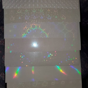 May include: A stack of eight iridescent holographic sheets with different patterns, including hearts, circles, stars, and lines. The sheets are white with a subtle shimmer.