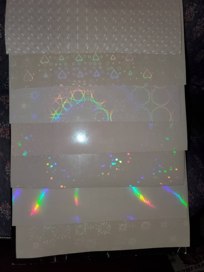 May include: A stack of eight iridescent holographic sheets with different patterns, including hearts, circles, stars, and lines. The sheets are white with a subtle shimmer.