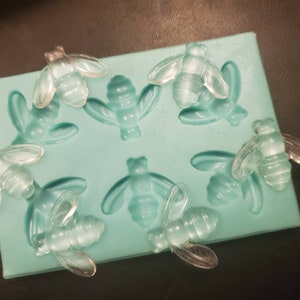 May include: A silicone mold with eight bee-shaped cavities. The mold is light blue and the bees are clear.