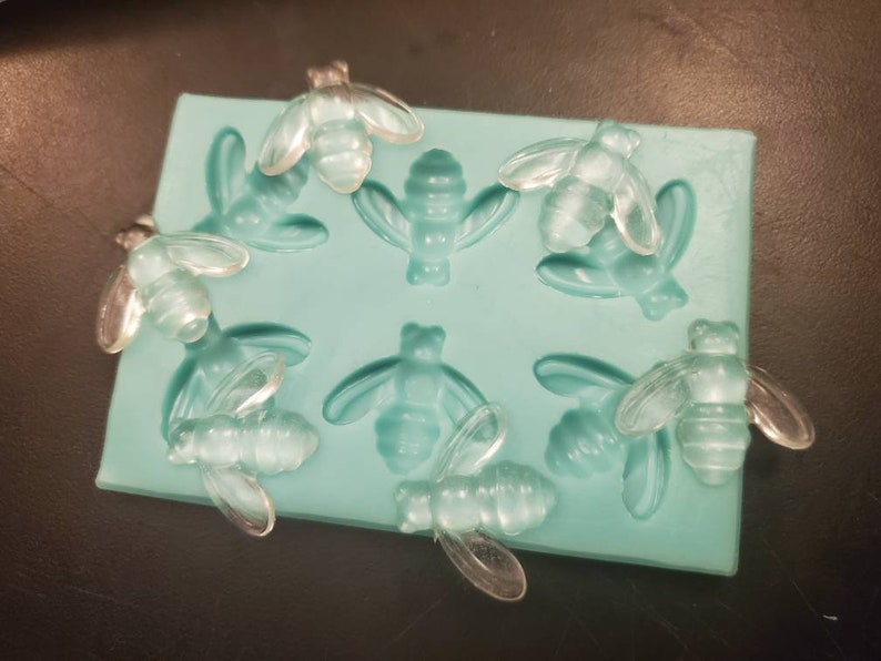 May include: A silicone mold with eight bee-shaped cavities. The mold is light blue and the bees are clear.