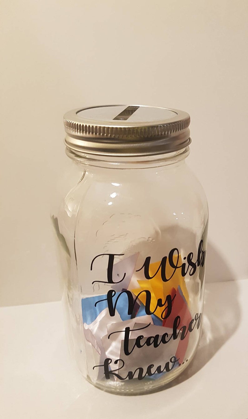 May include: A clear glass jar with a silver lid and a slot for inserting items. The jar has a black vinyl sticker that reads "I wish my teacher knew..." and is filled with colorful folded pieces of paper.