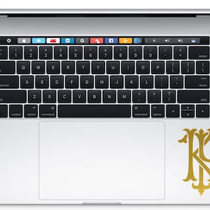 May include: A gold-colored monogram sticker with the letters "KP" in a decorative font, placed on the trackpad of a silver laptop.