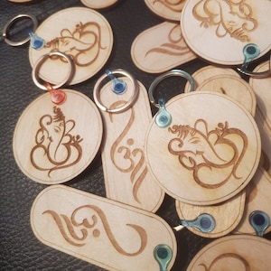 Puede incluir: Una colección de llaveros de madera con varios diseños, incluyendo la deidad hindú Ganesha y el símbolo sánscrito Om. Los llaveros están hechos de madera marrón claro y tienen un acabado natural. Cada llavero tiene un anillo de metal unido a él.