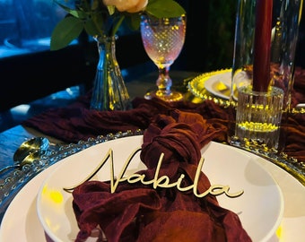 May include: A pink and white dinner plate with a burgundy napkin and a wooden name card that says "Nabila".
