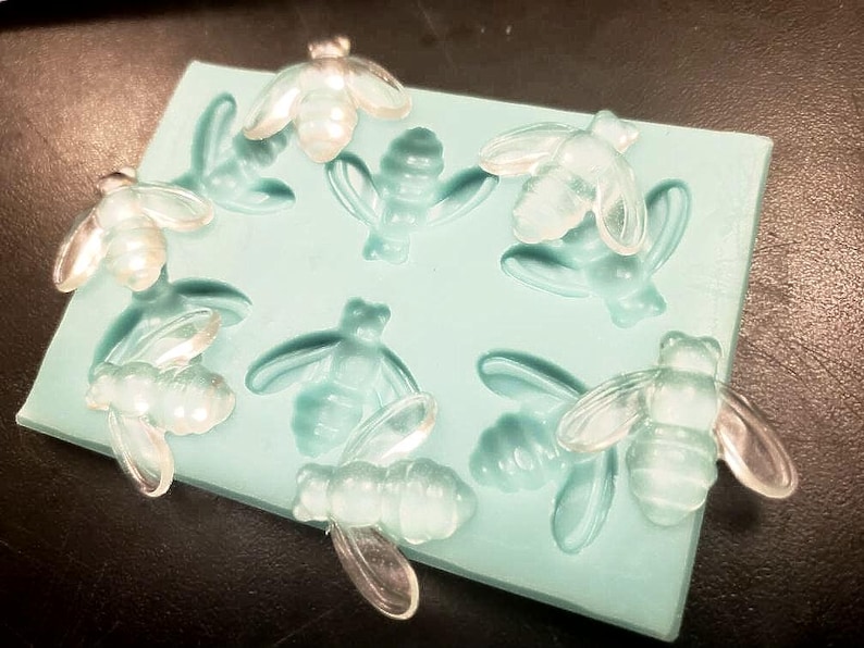 May include: A silicone mold with eight bee-shaped cavities. The mold is light blue and the bees are clear.