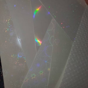 May include: A collection of iridescent holographic sheets with different patterns, including stars, circles, and fireworks. The sheets are translucent and have a rainbow sheen.