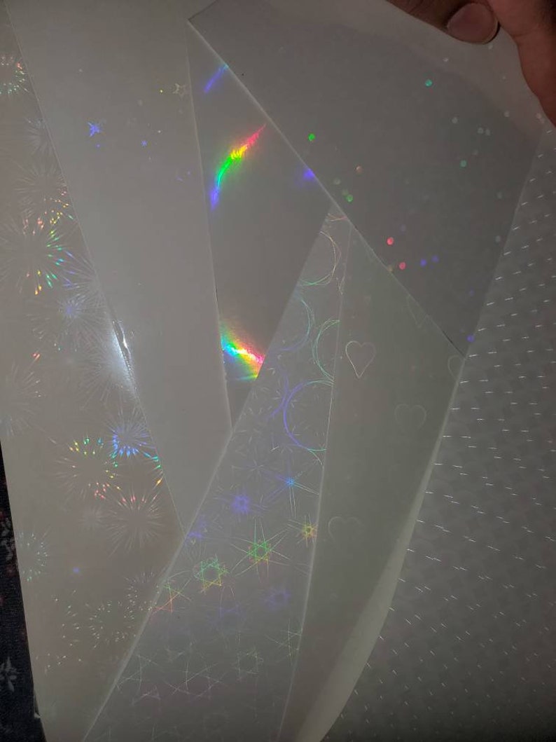 May include: A collection of iridescent holographic sheets with different patterns, including stars, circles, and fireworks. The sheets are translucent and have a rainbow sheen.