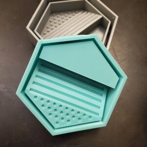 Op de afbeelding: Een tealblauwe siliconen mal in de vorm van een zeshoek met een kleinere zeshoek erin. De mal heeft een getextureerd oppervlak met verhoogde lijnen en stippen.