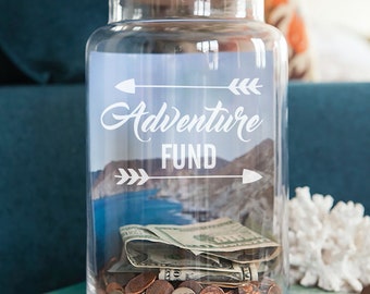 May include: A clear glass jar with a cork lid and a white sticker that says "Adventure Fund" with arrows on either side. The jar is filled with coins and bills.