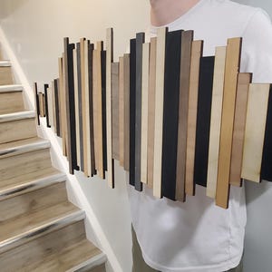 May include: A wall art piece made of wood strips in various shades of brown, black, and beige. The strips are arranged in a wave-like pattern, creating a modern and abstract design.