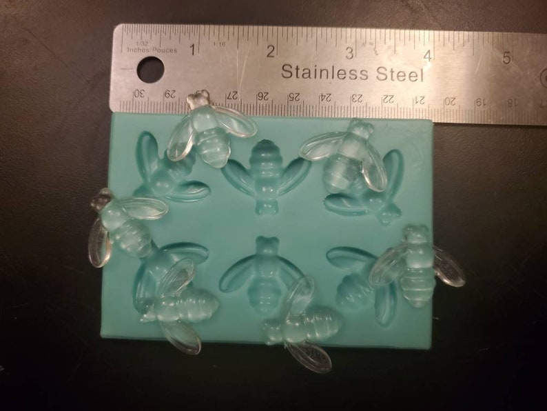 May include: A silicone mold with eight cavities shaped like bees. The mold is light blue and has a ruler with the words "Inches Pouces" and "Stainless Steel" printed on it.