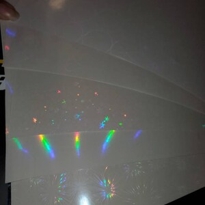 May include: A stack of four sheets of iridescent holographic paper with different patterns. The top sheet has a subtle shimmer, the second sheet has a pattern of stars, the third sheet has a pattern of vertical lines, and the bottom sheet has a pattern of fireworks.
