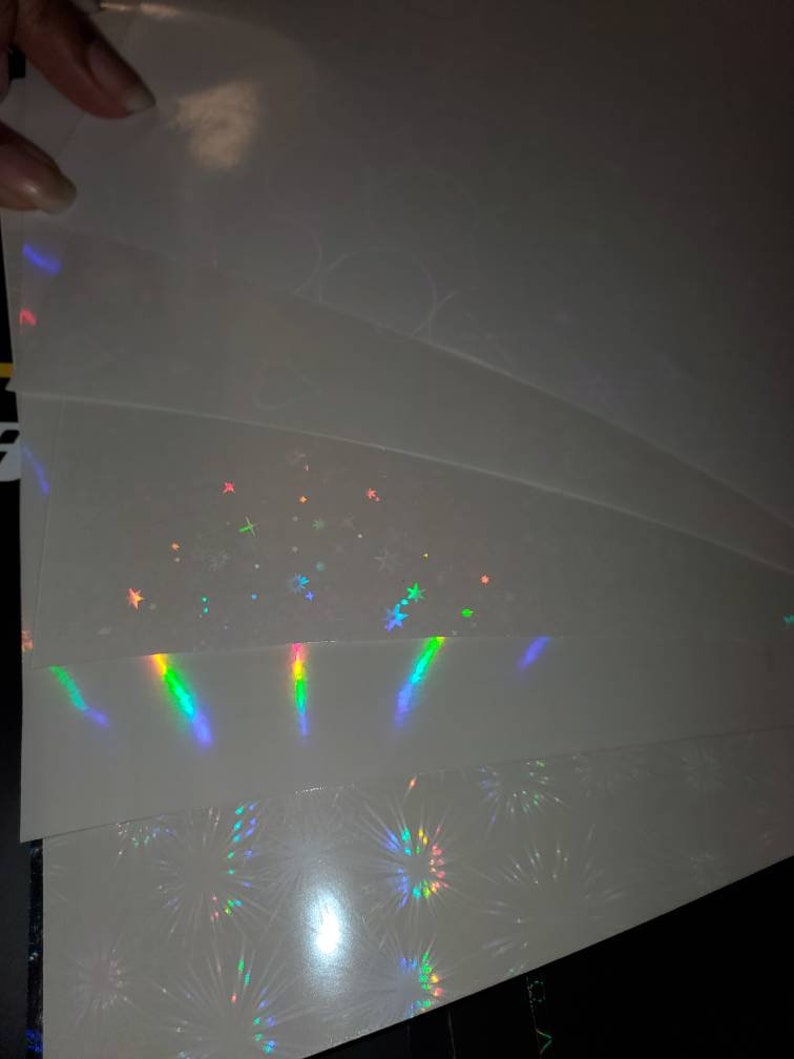 May include: A stack of four sheets of iridescent holographic paper with different patterns. The top sheet has a subtle shimmer, the second sheet has a pattern of stars, the third sheet has a pattern of vertical lines, and the bottom sheet has a pattern of fireworks.