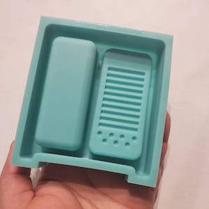 May include: A light blue silicone mold for making two phone-shaped soaps. The mold has a rectangular shape with two compartments. The left compartment is a simple rectangle, while the right compartment has a raised design of a phone with a speaker and buttons.