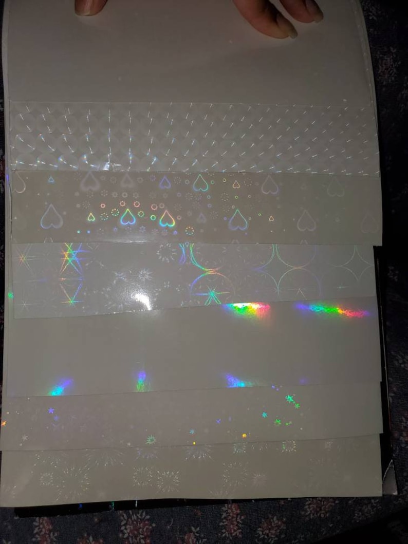 May include: A set of six iridescent holographic sheets with different patterns. The patterns include hearts, stars, and fireworks.