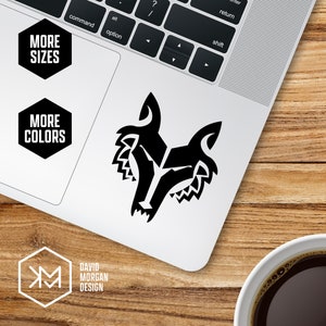 Könnte beinhalten: Ein schwarz-weißes Wolfskopf-Grafikdesign auf einem weißen Laptop. Der Laptop befindet sich auf einer Holzoberfläche. Auf dem Laptop befinden sich zwei schwarz-weiße sechseckige Aufkleber mit der Aufschrift "Mehr Größen" und "Mehr Farben".