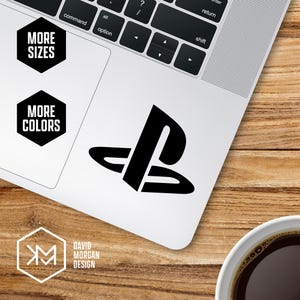 Puede incluir: Una pegatina negra con el logotipo de PlayStation en un portátil. El portátil está sobre una superficie de madera. La imagen también incluye el texto "MORE SIZES" y "MORE COLORS" en hexágonos negros, y un logotipo que dice "DAVID MORGAN DESIGN".