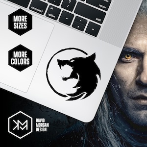 May include: Black and white sticker of a wolf's head inside a circle. The sticker is on a white laptop. The text "MORE SIZES" and "MORE COLORS" is on the laptop. The text "DAVID MORGAN DESIGN" is also on the laptop.