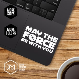 Puede incluir: Un portátil negro con una pegatina de vinilo blanco que dice "Que la fuerza te acompañe". La pegatina tiene una fuente inspirada en Star Wars y una estrella con dos estrellas más pequeñas dentro. El portátil está sobre una superficie de madera con una taza de café.