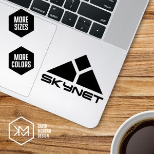 May include: A black and white Skynet sticker on a laptop. The sticker features a geometric design and the word "SKYNET" in bold letters. Additional text includes "MORE SIZES" and "MORE COLORS". The laptop is on a wooden surface.