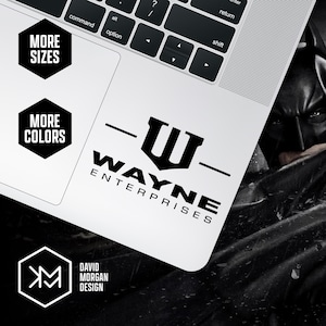 May include: A white laptop with a black and white Wayne Enterprises logo sticker. The sticker features the text "WAYNE ENTERPRISES" below a stylized "W" symbol. Additional black hexagonal graphics read "MORE SIZES" and "MORE COLORS".