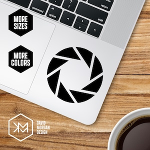 May include: A black aperture sticker is placed on a laptop. The sticker is a geometric design with triangular shapes. The laptop is on a wooden surface. The words "MORE SIZES" and "MORE COLORS" are on the laptop. The logo for "DAVID MORGAN DESIGN" is also visible.