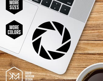 May include: A black aperture sticker is placed on a laptop. The sticker is a geometric design with triangular shapes. The laptop is on a wooden surface. The words "MORE SIZES" and "MORE COLORS" are on the laptop. The logo for "DAVID MORGAN DESIGN" is also visible.