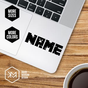 May include: A white laptop with a black pixelated "NAME" sticker. The laptop also has two black hexagon stickers that say "MORE SIZES" and "MORE COLORS". A coffee cup is in the bottom right corner.