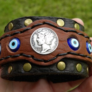 May include: A brown leather bracelet with a silver coin, a black leather band, and blue evil eye charms. The coin has the words "In God We Trust" and "1944" on it.