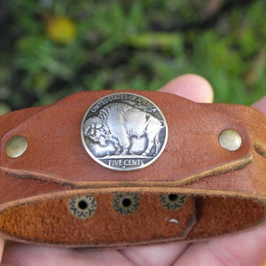 Op de afbeelding: Een bruin leren armband met een zilveren muntstuk van vijf cent met een buffel en de tekst "United States of America" en "Five Cents".