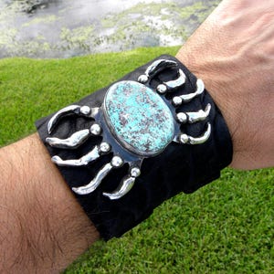May include: A black leather bracelet with a silver claw design and a turquoise stone in the center.