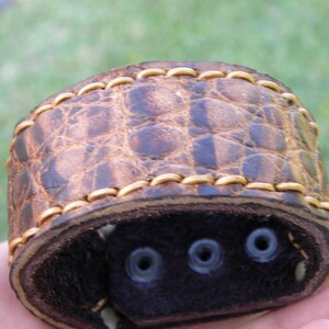 May include: A brown leather watch band with a crocodile pattern and gold stitching. The band has three holes for adjusting the size.