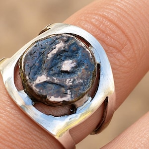 May include: A silver ring featuring a circular, weathered, dark brown and blue metallic coin set in the center. The band is smooth and polished, with open cutouts on the sides that frame the coin, creating a contrast between the shiny metal and the aged, textured surface of the coin.
