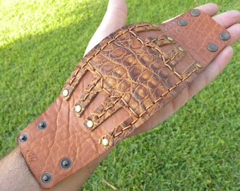 May include: A brown leather bracelet with a crocodile-embossed design and brass rivets. The bracelet has a cut-out pattern and is stitched with brown thread.