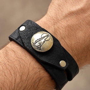 May include: A black leather cuff bracelet featuring a textured surface and a circular silver-toned metal snap button. The button is embossed with a detailed graphic illustration of a wolf howling at a full moon, secured by two silver metal rivets.